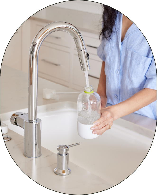 A person standing at a running sink, adding water to the water fill-line of their reusable Glass All-Purpose bottle. - Card 1