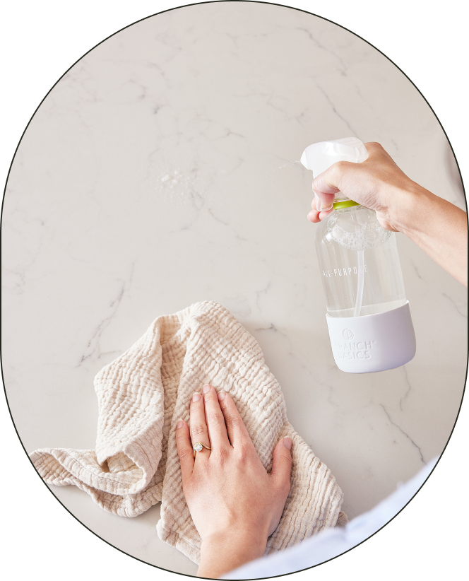 A person using an All-Purpose bottle of Branch Basics Concentrate in their right hand to spray a white marble surface; they are using a lint-free rag in their opposite hand to wipe the surface. - Card 3