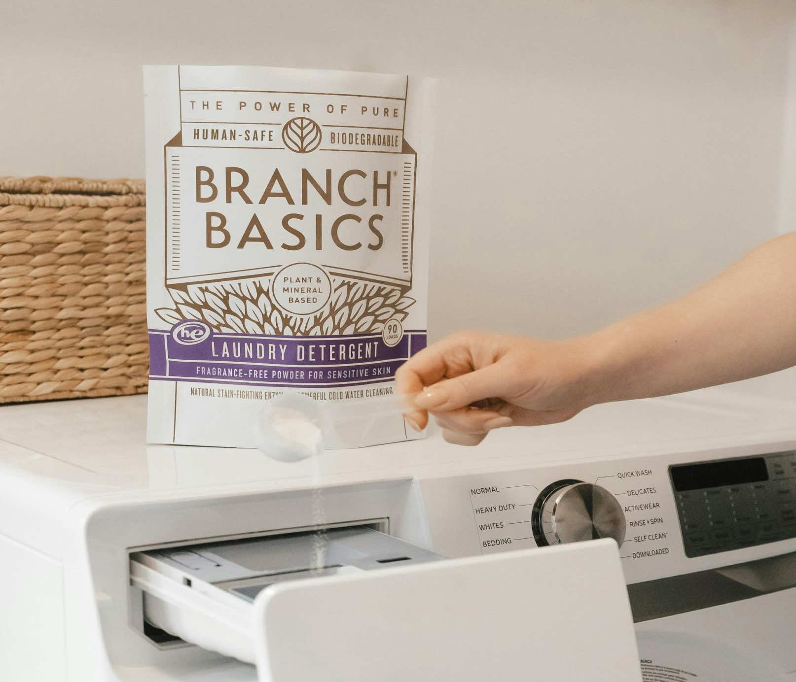A bag of Laundry Detergent on top of a washing machine, with a scoop of detergent being added to the washing machine. - View 8
