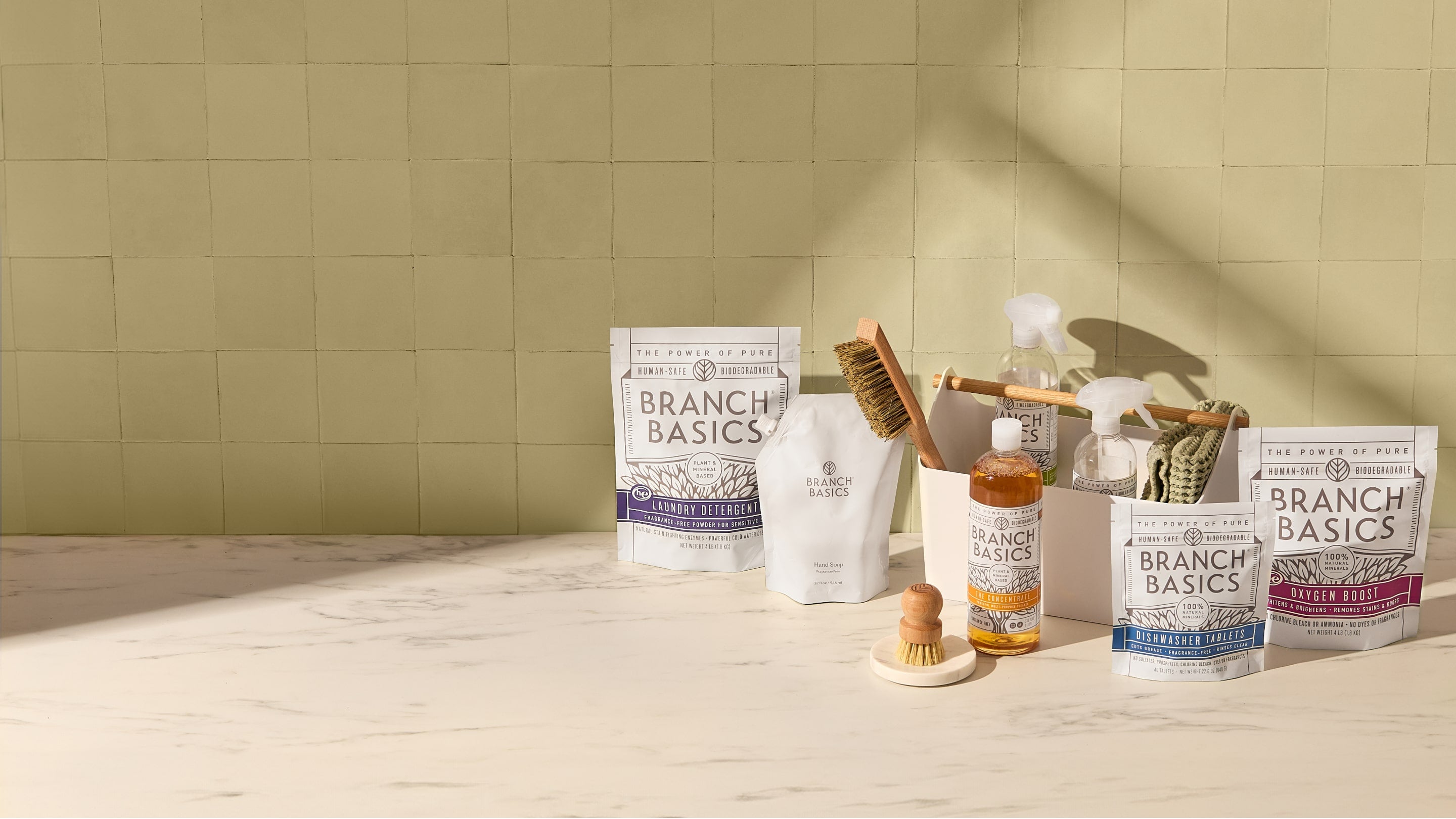 Various Branch Basics cleaning products, including spray bottles, a brush, and packages, are arranged on a light-colored countertop against a tiled wall.