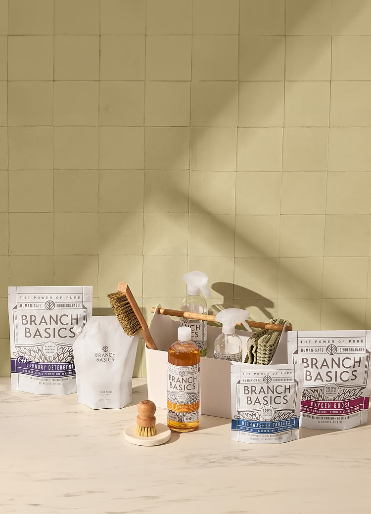 Various Branch Basics cleaning products, including spray bottles, a brush, and packages, are arranged on a light-colored countertop against a tiled wall.