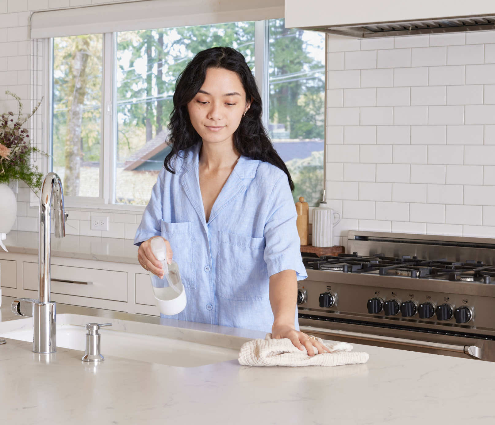 A woman standing at a kitchen counter, using a Glass Branch Basics bottle and a cloth towel to clean the counter's surface. - View 6
