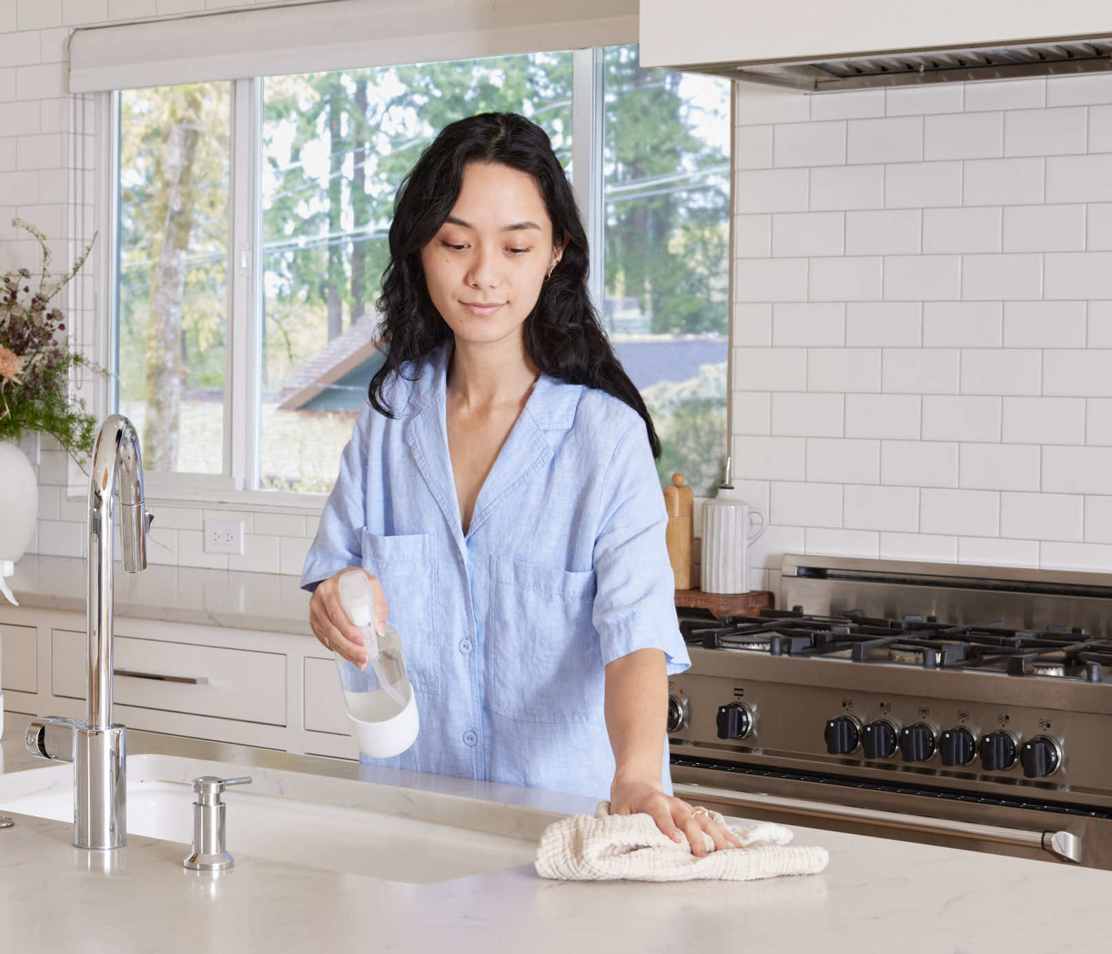 A woman standing at a kitchen counter, using a Glass Branch Basics bottle and a cloth towel to clean the counter's surface. - View 3

