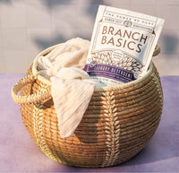 Wicker basket with folded beige towel and Branch Basics laundry detergent packet on a light purple surface. - Thumbnail View 5
