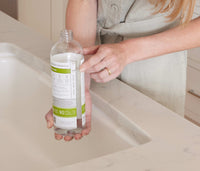 A woman standing at a kitchen faucet, pointing at the fill-lines on a refillable Plastic Branch Basics bottle; there are separate fill-lines for water and Branch Basics Concentrate, respectively. - Thumbnail View 3
