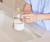 A woman standing at a kitchen faucet, pointing at the fill-lines on a refillable Glass Branch Basics bottle; there are separate fill-lines for water and Branch Basics Concentrate, respectively. - Thumbnail View 4
