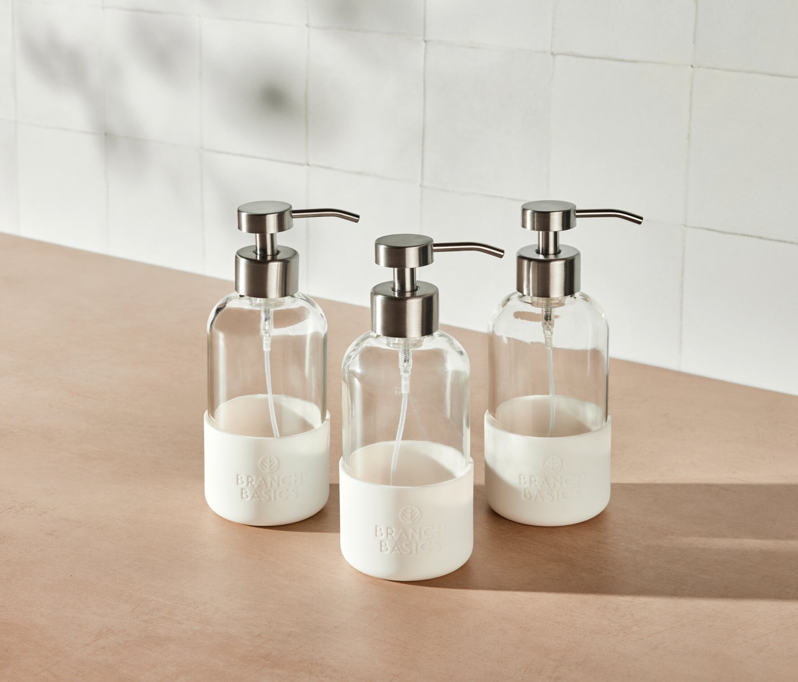 A trio of refillable Glass Foaming Wash Bottles with Brushed-Chrome pumps, displayed on a neutral surface and white tiled background. - View 2
