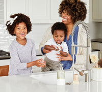 A single refillable Glass Foaming Wash Bottle with a Brushed-Chrome pump being used by a mom and two children to wash their hands in a kitchen sink. - Thumbnail View 3
