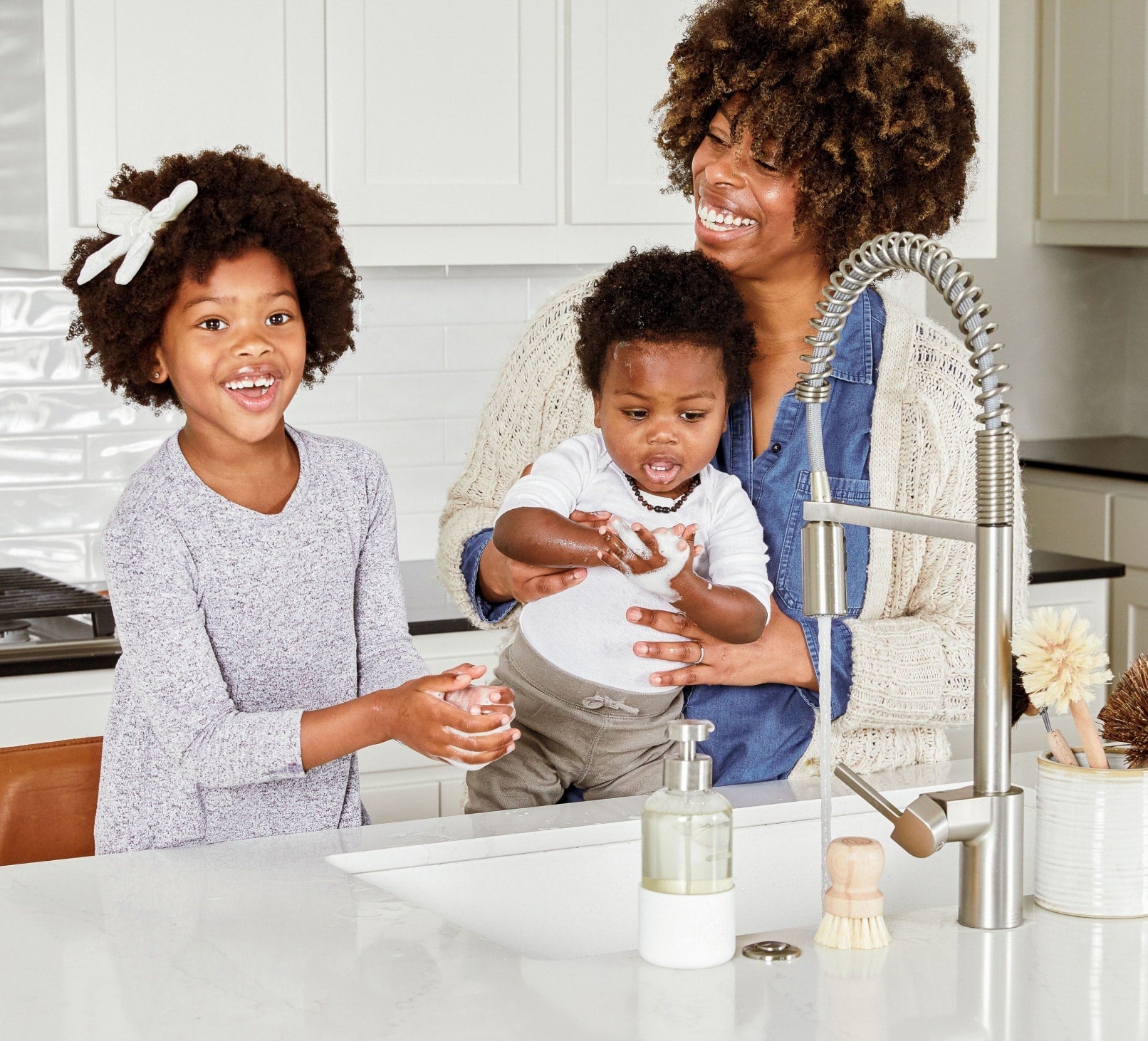 A single refillable Glass Foaming Wash Bottle with a Brushed-Chrome pump being used by a mom and two children to wash their hands in a kitchen sink. - View 3
