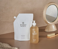 A pouch of Branch Basics Gel Hand soap standing next to a full Glass Bottle of Gel Hand Soap, displayed on a neutral background. - Thumbnail View 4
