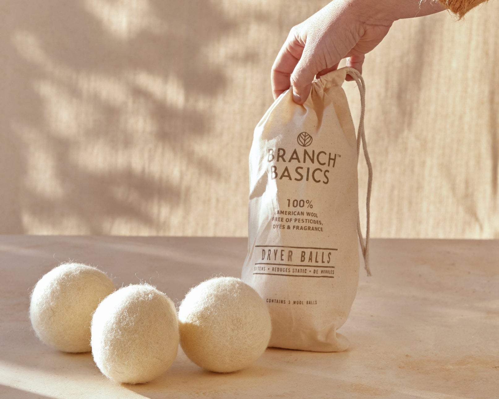 A trio of Wool Dryer Balls accompanied by a cloth carrying bag that is being held up by a person's hand, displayed on a neutral surface and neutral background. - View 4
