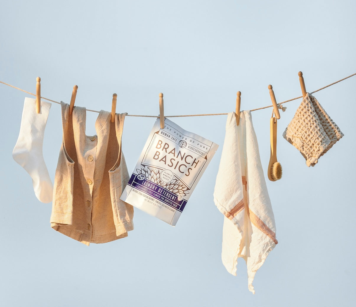 Clothing items hanging on a clothesline with Branch Basics Laundry Detergent packaging against a clear blue sky. - View 4
