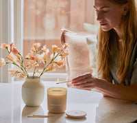 A person uses a lit match to ignite the 100% Beeswax Candle as it sits on a stone tabletop. - Thumbnail View 5
