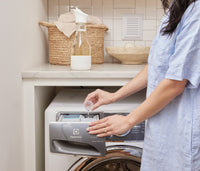 A person pouring diluted Branch Basics Concentrate from the lid of a refillable Glass Laundry Bottle into the detergent compartment of a washing machine. - Thumbnail View 3
