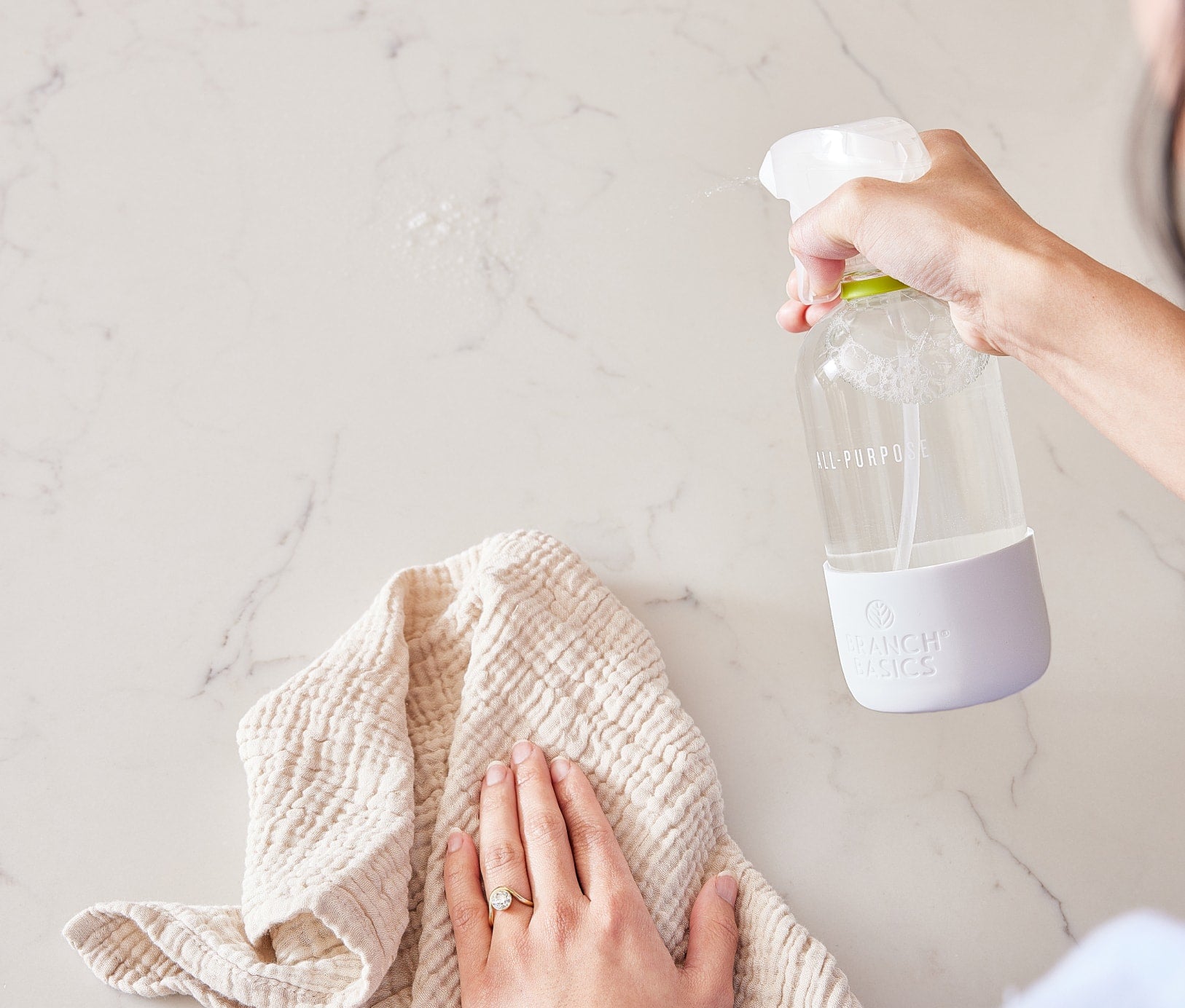 A person is holding a Glass All-Purpose Spray bottle in one hand and a cloth hand towel in the other, using both to clean a stone countertop. - View 3
