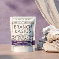 A pouch of Branch Basics Laundry Detergent on a wooden surface with a purple background. - Thumbnail View 1
