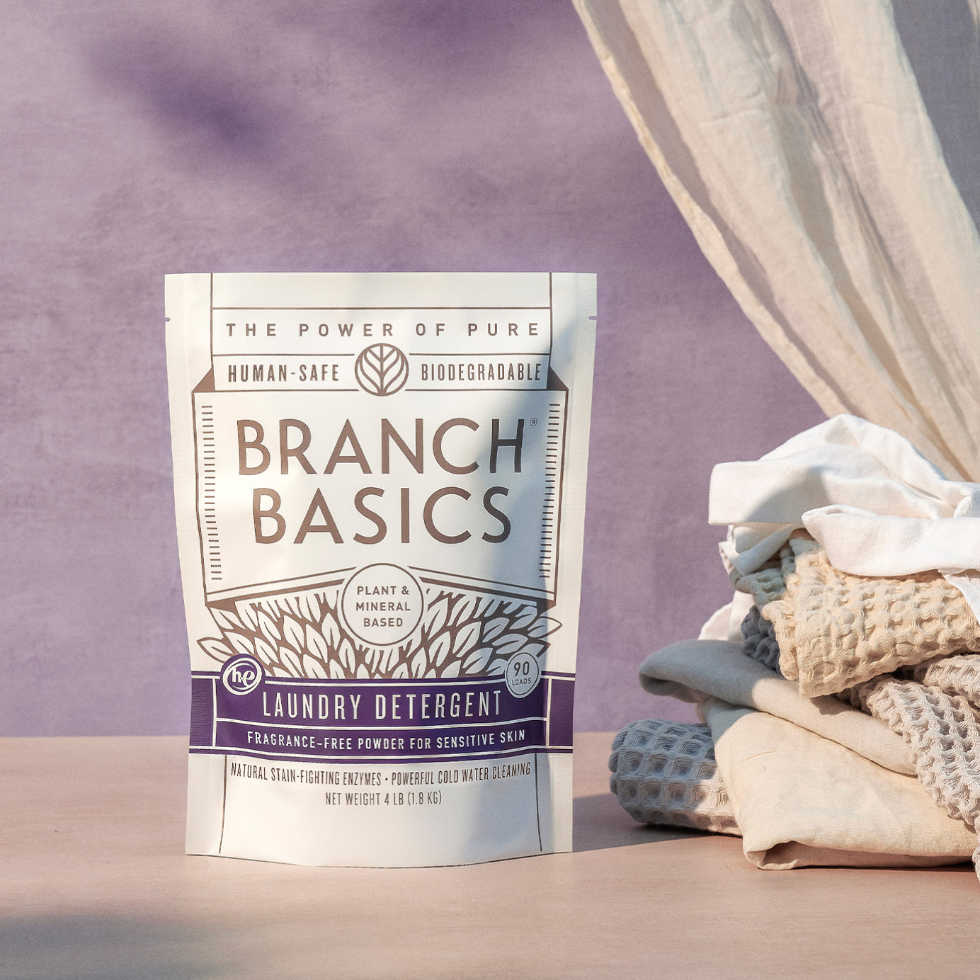 A pouch of Branch Basics Laundry Detergent on a wooden surface with a purple background. - View 1
