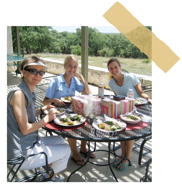 Marilee, Allison, and Kelly sitting at an outdoor table with food and drinks, smiling; image contained within a frame to emulate the look of a polaroid.