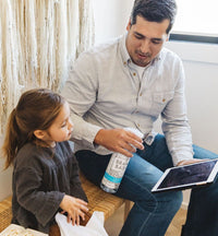 An adult and child sitting together on a bench, using a Plastic Streak-Free bottle to clean fingerprints from the glass screen of a tablet device. - Thumbnail View 3
